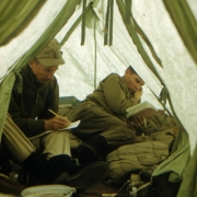 View of Captain Albert Jackman and Lieutenant Paul Townsend of the 87th Mountain Infantry Regiment (re-enforced) inside a green Everest Tent during the Mount Rainier Test Expedition. Jackman is taking notes and Townsend is reading. Equipment includes a stove, boots and crampons, and a sleeping bag. Jackman is wearing a cap, a dark shirt, khaki pants, and smooth-soled black boots. Townsend is wearing khaki shirt and pants and smooth-soled black boots.