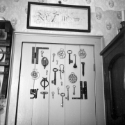 Keys of many different shapes hang on the back of a paneled door. Parts of furniture are visible of either sides of the door. There is a framed piece of embroidery with a cross, a crown, and "No no" hanging on the wall. The wallpaper is patterned with rural scenes surrounded by ornate decorative patterns.