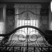 A  double bed in the Prosser house is illuminated by the light streaming in through the lace curtains of the window behind it. The brass headboard against the window is decorated with spindles and curved brass. The brass footboard is arched and has decorative brass pieces inside it. A quilted spread is on the bed.