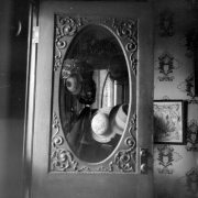 Straw and cloth hats hanging on a hatrack are reflected in an oval mirror in the Prosser house. Sunlight illuminates the left side of the image. The mirror has a rectangular frame and scroll-work around the edges. Another mirror is visible in the reflection of the mirror. An embroidered, framed picture hangs to the right. The wallpaper has a decorative pattern on it.