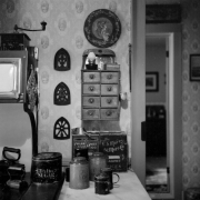 A stove sits next to a marble-topped cabinet in a kitchen in Central City, Colorado. The stove has an oven and a warmer with enamel doors. Two irons are on the range, and an assortment of bric-a-brac, such as a coffee grinder and plates, is on top of the stove. A sugar tin is next to the stove. Staple tins and utensils are on the counter. A small chest with ten drawers is next to an open doorway. Decorative items hang on the walls. A picture is on the wall in the background.
