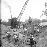 View of (probably) Lipan Street in Denver, Colorado; shows a ditch with shoring, and men working with a clam shovel crane.