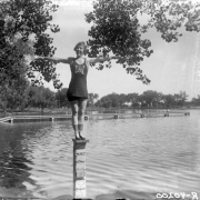 A young woman poses on a pillar in Smith Lake at Washington Park, Denver, Colorado. A sign on the nearby boardwalk over the water reads: "Women Only Do Not Go Out In The Lake Beyond The Depth Of Your [?]".