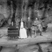 Four musicians (two women and two men) pose for a photo at Red Rocks Amphitheater, Morrison, Colorado, near a sounding board. One man holds a cello, a woman sits at a portable piano, and two singers stand Wooden planks are on the ground in the foreground.