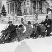 Men, children and a woman pose on a sled near the David Moffat mansion at 808 Grant Street in Denver, Colorado; shows Bert Rhoads (1st on sled), Charles Gates (waving), Hazel Rhoads Gates (holding onto her hat) and Harry Fisher Rhoads (holding one of the Gates' daughters).