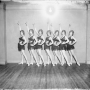 Dancers from the Georgia Land School of Dancing pose for a group photo in Denver, Colorado. The women, part of the Georgia Lane Group, wear short fringed skirts, bikini tops with sheer fabric overlays, and  have their hair marcelled (waved). The woman posed at the extreme left is Sarah Francis "Sally" Orr.