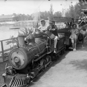 Young women passengers aboard the miniature steam train at Lakeside Amusement Park in Lakeside, Colorado, wave to the photographer. A young man and woman sit in the engine of the train as the man pulls the cord to blow the whistle. Shows a roller coaster and park building in background.