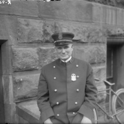 Captain Daniel Cronin poses near a stone building.  He wears a uniform with brass buttons down the front, a badge, hat and four bars on his collar. A bicycle leans against the building.