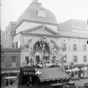 An exterior view of the Orpheum Theater, a vaudeville theater, located on 1537 Welton Street in Denver, Colorado. The theater is decorated with ribbons and small international flags for All Nationality Week. A sign reads: "Orpheu[m] Modern Vaudevi[lle]." The Orpheum Theater opened in 1903, was razed in 1930 then rebuilt two years later.