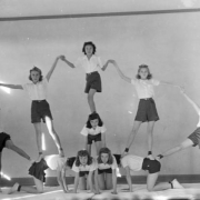An eighth grade girl's gym class forms a human pyramid at the Central School in Alamosa, Colorado. The girls wear white, short sleeved blouses, belted dark shorts, socks and white tennis shoes. Four girls are on their hands and knees in the center. Another girl is on her hands and knees on the top center of the four girls. Another stands on her back, with others standing, holding hands with outstretched arms, to form a large arch that spreads across the room.