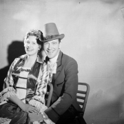 A man and a woman dressed in costume for Saint Patrick's Day pose, probably Denver, Colorado. She wears a dress with ruffled sleeves, lace and satin bodice, and a tiara. He wears a hat and suit with satin and wide lapels.