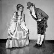 A man and a woman dressed in costume for Saint Patrick's day pose for a photo. She wears a long patterned dress with ruffled sleeves, lace and satin bodice, and a tiara. He wears a hat, suit with satin and wide lapels, knickers and tights.