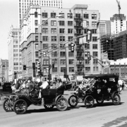View of the Saint Patrick's Day Parade at 16th (Sixteenth) Street and Broadway in the Central Business District, Denver, Colorado. Men and a woman in hats ride in decorated antique convertible cars. The Paramount Theater and Daniels and Fisher Tower are in the distance.