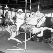 A young girl rides a carousel horse on the merry-go-round at the Lakeside Amusement Park in Denver, Colorado. The child wears a white pants and blouse, button shoes, and a hat.