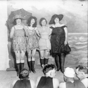 Four women in bathing attire (bathing suits), one with an umbrella, pose at a Gates Company costume party in Denver, Colorado. Mrs. Hazel Gates is on the far right in black.