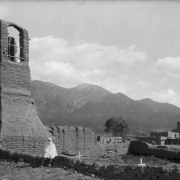 View of mission ruins and graves at Taos, New Mexico; a Native American (Taos Pueblo) poses in a blanket. Adobe pueblo dwellings and Pueblo Peak are in the background.