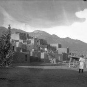 View of Taos Pueblo, New Mexico; shows a Native American (Taos Pueblo) man, scaffolds, adobe pueblo dwellings, and drying laundry.