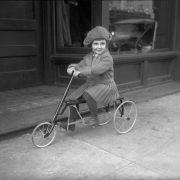 Mary Elizabeth Rhoads, daughter of Harry M. Rhoads, rides a Speed Flivver tricycle in Denver, Colorado. She wears a wool dress and hat.