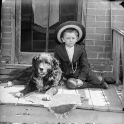 Outdoor portrait of a boy and his dog on the porch of a house in Denver, Colorado. The boy wears knickers and a wide-brimmed hat.