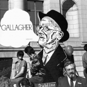View of the James Joyce Society float in the Saint Patrick's Day parade stopped in front of the Denver City and County Building, Civic Center, Denver, Colorado. The float has a cut-out sign of a profile of James Joyce that reads: "Gallagher, Ulysses, page 135." A political sign in the distance reads: "Gallagher [for] Mayor." State Senator Dennis Gallagher is standing in front of the float.