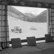 View (from inside) of snow, a mountain, and a tracklayer vehicle, probably in Colorado. Interior furnishings include a ledge seat, pillows, and a flower patterned valance.