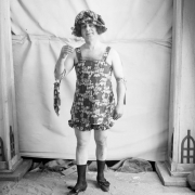 Photographer Harry M. Rhoads poses in costume. He wears a floral patterned women's bathing suit with a matching bonnet and hand bag, women's shoes and black socks.