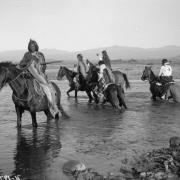 View of a Native American (Ute) scouting party mounted on horseback as they cross the Los Pinos river, La Plata County, Colorado; three men have rifles, one a pistol; men wear moccasins, fringed leggings, blankets, shirts, and braided hair; some have feathers in their hair; all horses have bridles and saddles.
