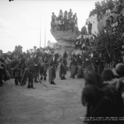 Native American men (Hopi), snake dancers, stand in a circle at Walpi Pueblo, First Mesa, Arizona; five men handle snakes, one holds a snake in his mouth; one man strokes a rattle snake (on the ground) with a feather, while groups of unidentified Native American and white men and women look on; snake members wear kilts and bead necklaces; some wear body paint.