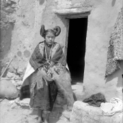 Outdoor portrait (standing) of a Native American (Hopi) woman near a Pueblo doorway at Walpi Pueblo, First Mesa, Arizona; she wears a dark manta, print and fringed shawl, woven blanket shawl, bead necklace with open hoop pendant, and hair in side whirls.