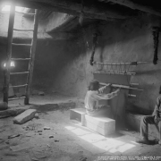 Interior view of a kiva, possibly the snake kiva, at Walpi Pueblo, First Mesa, Arizona; two unidentified Native American (Hopi) men sit near a loom; a wooden ladder leans against a large opening in the ceiling; a clay bowl is next to the ladder; one man sits on a wooden crate at a wooden loom suspended from the ceiling by fur pelts, and weaves a dark blanket or rug; one man sits against the wall; both men wear pants and print shirts; one wears no shoes.