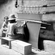 Interior view of a kiva, possibly the snake kiva, at Walpi Pueblo, First Mesa,  Arizona; two unidentified Native American (Hopi) men sit in the kiva; one man sits on a wooden crate at a wooden loom and weaves a dark blanket or rug while another looks on; both men wear pants and print shirts, one wears a woven hat, one wears a headband and no shoes.