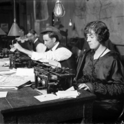 Rocky Mountain News reporters (men and women) work at a table with typewriters in Denver, Colorado; newspapers and notes are piled on the table.