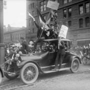 World War One military recruits ride a car in a Denver, Colorado, parade. The men hold signs, United States flags, blow party horns and wave to the crowd. Picket sign reads: "The Kaiser Has;" a dead duck hangs from it. Office buildings are in the background.