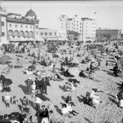 People with umbrellas crowd Santa Monica Beach in Santa Monica (Los Angeles County) California. Shows businesses and the Ocean Park Bath House, a Moorish style building with arches and domes.