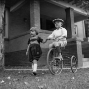 Charles C. Gates Jr. rides a tricycle in a Denver, Colorado yard while one of his sisters walks along side him. He wears shorts, shirt and a cap. His sister wears a knee length dress.