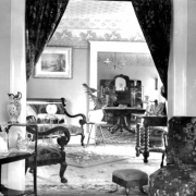 Interior view of a Denver, Colorado residence; decor includes brocade upholstery, a settee, chairs, tables, a foot stool, china pitchers, crystal, carpet, rugs, and a potted palm.