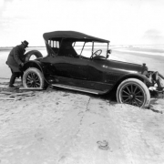 View of a car mired in mud by a river, (probably) in Colorado; a man lifts the spare tire from the back of the vehicle.
