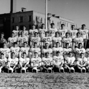 Portrait of a football team in Trinidad, Colorado. Teenage boys and young men in white, numbered jerseys are posed, the ones sitting in the front have their helmets on the grass in front of their cleated shoes. Two men in ballcaps and jackets with large "T" patches flank the standing row. Power poles and a three-story, flat roofed cream colored brick building is in the background.