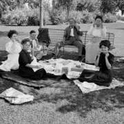A family picnic in an unidentified park, Colorado, includes, "from left to right: Mrs. Knapp (Julia's mother), Hazel (Rhoads) Gates, Charles C. Gates, Harry F., Addie, and Julia (Knapp) Rhoads." Each person holds a corn cob to their mouth.