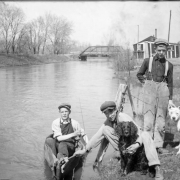 Outdoor portrait of boys with their dogs by Archer Canal in Denver, Colorado. One boy is in a canoe. Charles S. Lillybridge's house and studio and the Alameda Avenue bridge are in the distance.