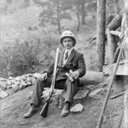 Photographer Harry Rhoads sits on a rock in a suit and pith helmet; he holds binoculars and a rifle; a pistol is in his jacket pocket, probably Colorado.