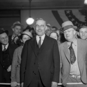 Denver Mayor Benjamin Franklin Stapleton (1873-1950) poses (eyeglasses and spotted tie), with a crowd of men and women, in Denver, Colorado; bunting is on the wall.