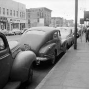 View of Welton Street in Denver, Colorado; shows parked cars, pedestrians, and business signs: "Jim Furlong Loans," "Baker Hotel," and "Estes Hotel."
