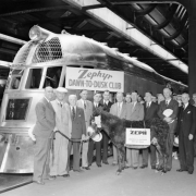 Men, boys, and girls pose with a burro in Chicago (Cook County), Illinois; and signs: "Zephyr Dawn to Dusk Club" and "ZEPH Denver to Chicago May 26, 1934." A Chicago, Burlington, and Quincy Railroad locomotive is in the background. Fred G. Gurley, later president of the Atchison, Topeka and Santa Fe, is fourth from the right, with the wire rim glasses.