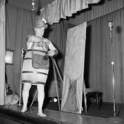 Portrait of photographer Harry M. Rhoads, on stage in (probably) Denver, Colorado, wearing a wooden barrel, a Fedora hat, and camera equipment bags. A microphone is to the side.