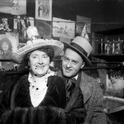 A man and woman pose. The woman wears a fur muff and fancy hat. Darkroom equipment and photographs are on interior walls.