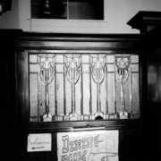 Interior view of a leaded glass window in the Columbian Hotel, in Trinidad, Colorado. Vertical panels of "ice" frosted glass contain leaf shapes interlocked with stirrup shapes at the top; a border with arches at regular intervals crosses the bottom. Signs under the window read: "Women" and "Dining Room".  Signs are illuminated by a shaded gallery lamp mounted on the polished wood frame that holds the window.