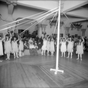 At the Gates Rubber Company's May Day celebration, several women in matching costumes, with headbands and ballet shoes, hold long ribbons attached to a Maypole. The May Day queen sits on a stage with a crown and a bouquet of flowers; John Gates stands behind her to the right. Shows interior with wall planters, sectional ceiling, and wood floor, Denver, Colorado.