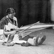 We'wha, a Native American (Zuni), sits and weaves a belt on a backstrap loom, Zuni Pueblo, New Mexico. We'wha is a berdache, a man who prefers women's work and adopts female dress; he is dressed as a woman and wears a woven manta, moccasins, and squash blossom necklace.