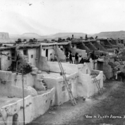 View looking east over Acoma Pueblo, Acoma Mesa, New Mexico, shows Pueblo Native Americans, multi-story adobe house blocks with ladders, vigas, chimneys, pottery, chickens, firewood and surrounding buttes and mesas.